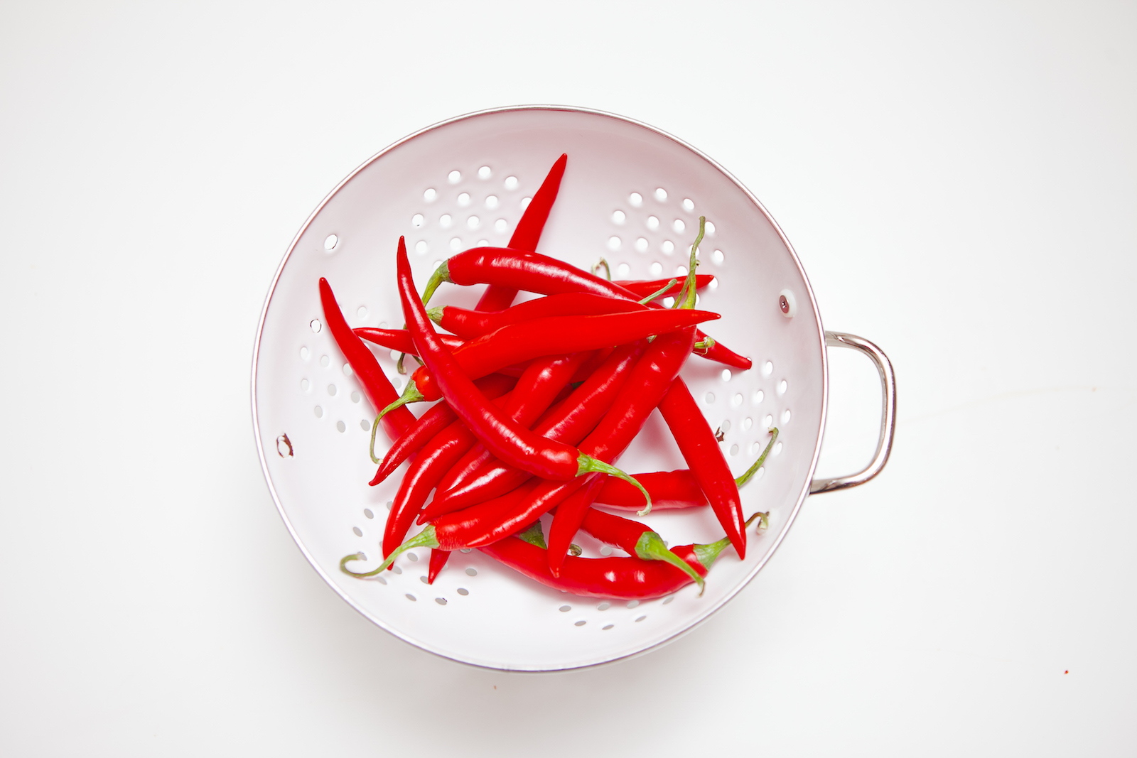 White colander with whole chillis in it against a white background